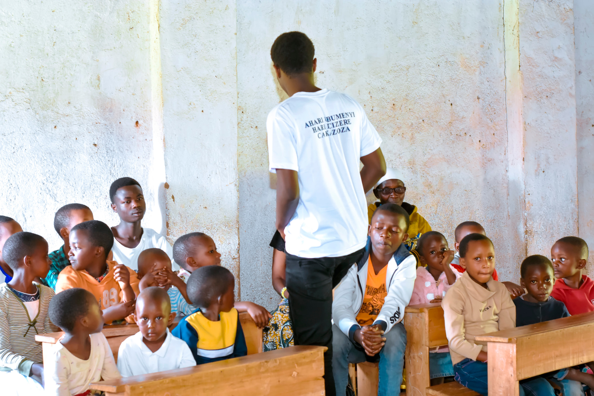 A volunteer speaking to a full classroom, wearing the shirt that reads “Ahari ubumenyi hari icizere ca kazoza”.